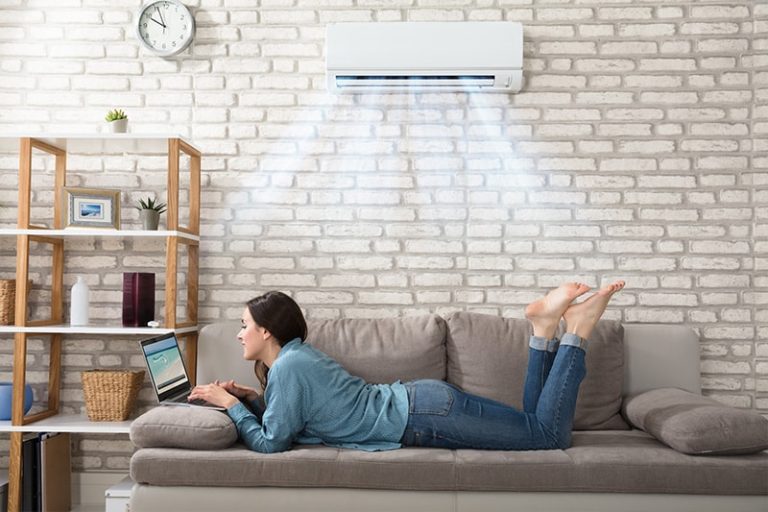 Woman Lying On Sofa Using Laptop Enjoying The Cooling Of Air Conditioner At Home